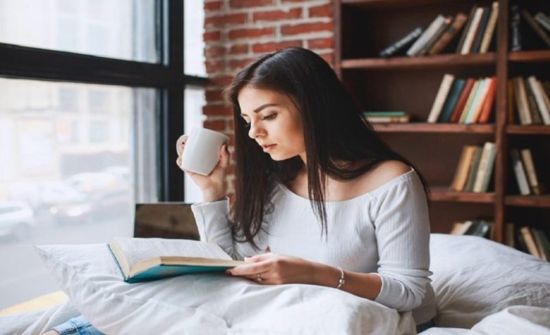 Perempuan Membaca Buku Di Atas Tempat Tidur Dengan Rak Buku Di Belakang
