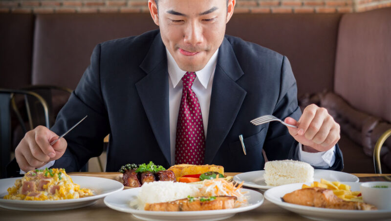 Young Asian Businessman In Formal Suit Feeling Hungry, Eating Many Foods On Table At Cafe